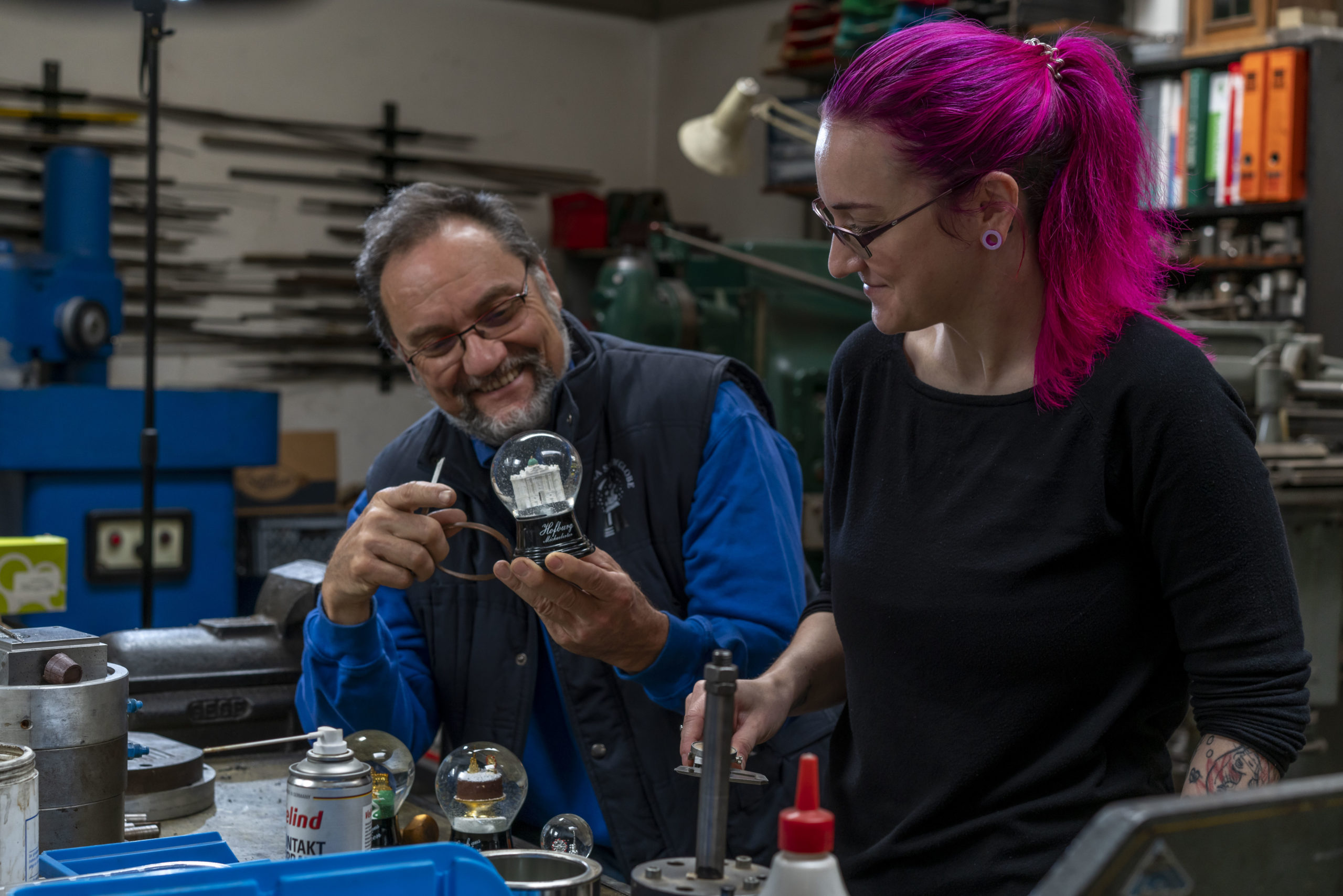 Zwei Personen betrachten in einer Werkstatt eine Schneekugel. Sie sind umgeben von Werkzeugen und Geräten.