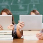 Zwei Schüler benutzen Tablets, die auf Bücherstapeln vor einer Tafel liegen.