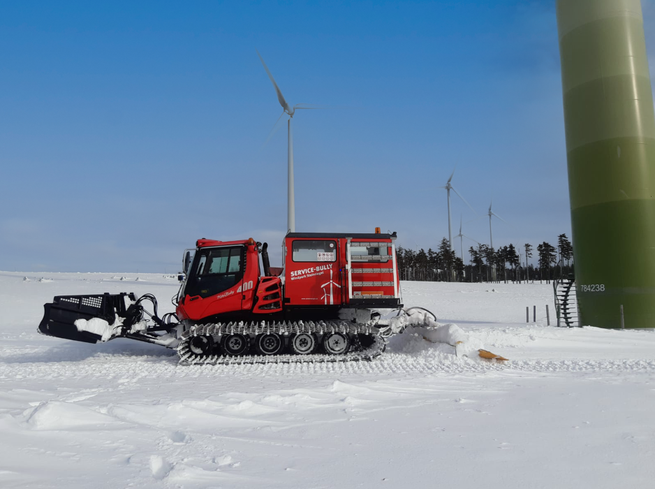 Ein rotes Pistenfahrzeug auf einem schneebedeckten Feld in der Nähe von den Windkraftanlagen Steinriegel und einem großen grünen Turm.