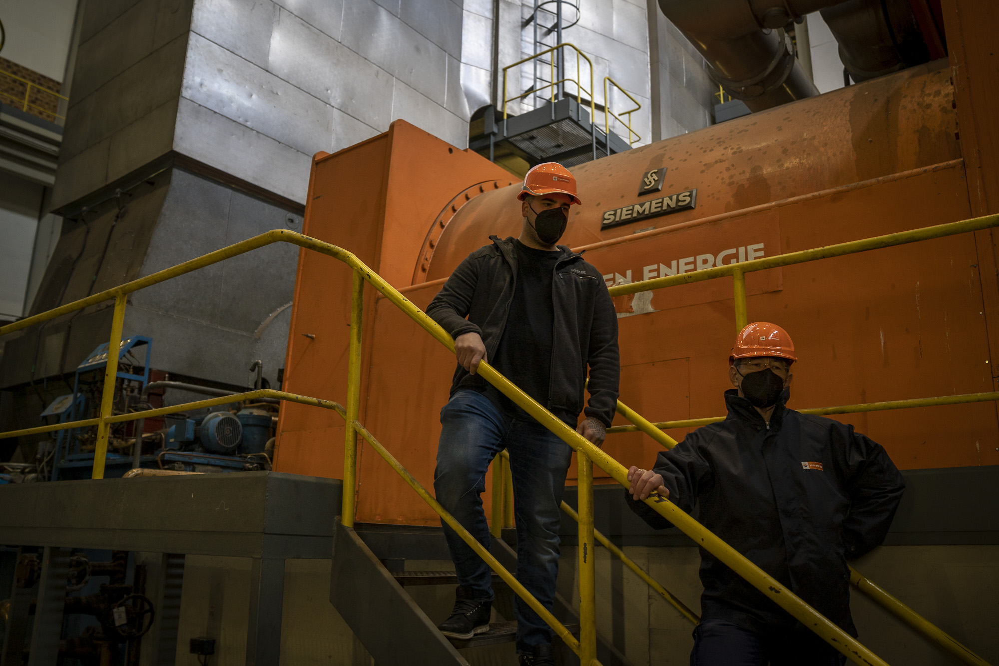 Zwei Arbeiter mit orangefarbenen Helmen und Masken stehen auf einer Treppe im Kraftwerk Simmering.