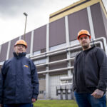 Zwei Personen mit orangefarbenen Schutzhelmen stehen vor einem Industriegebäude, dem Kraftwerk Simmering. Im Hintergrund ist ein Schornstein zu sehen.