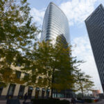 Modernes Stadtbild mit hohem, geschwungenem Glasgebäude, schrägem Turm, Bäumen und Menschen, die auf dem TownTown Platz spazieren gehen.