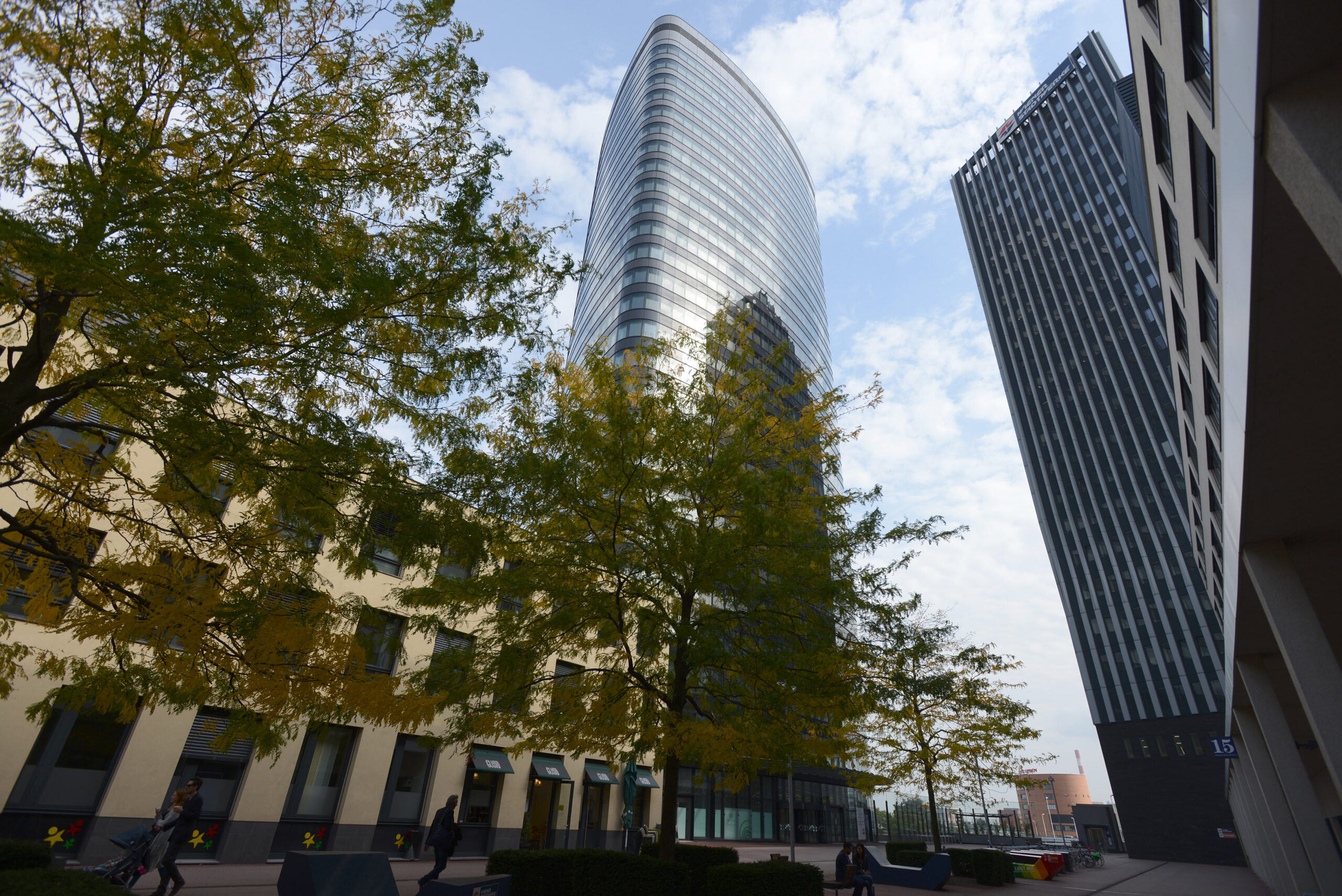 Modernes Stadtbild mit hohem, geschwungenem Glasgebäude, schrägem Turm, Bäumen und Menschen, die auf dem TownTown Platz spazieren gehen.