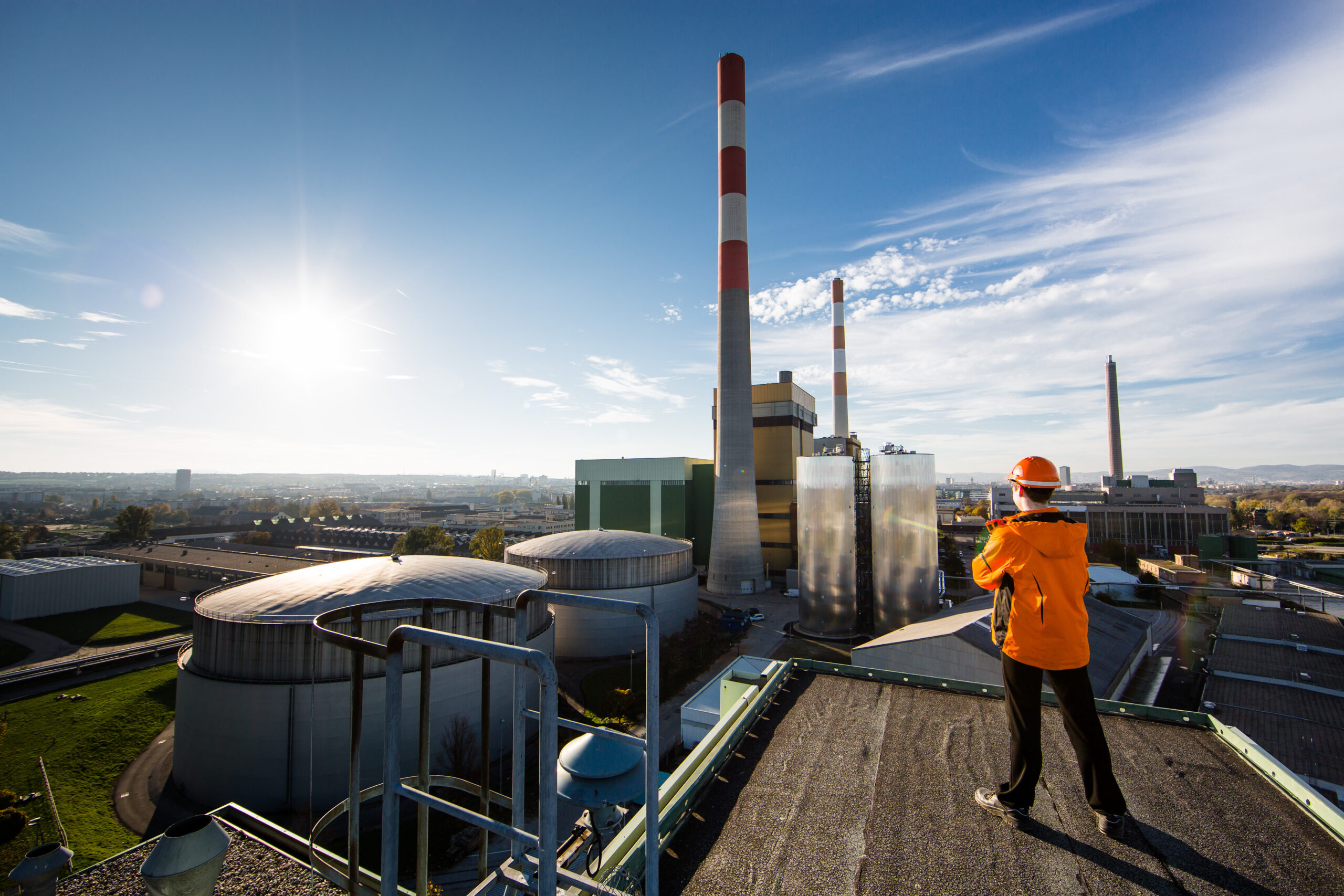 Person in orangefarbener Schutzausrüstung auf einem Dach mit Blick auf das Kraftwerk Simmering mit Schornsteinen unter einem sonnigen Himmel.