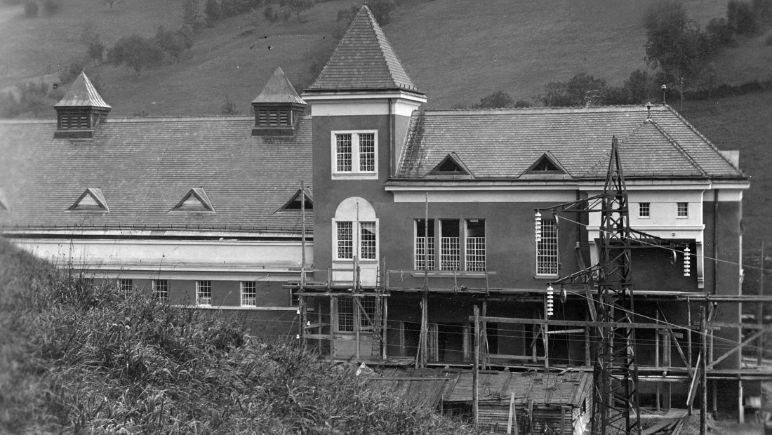 Ein großes im Bau befindliches Gebäude, dem Kraftwerk Opponitz, mit Gerüst, spitzen Dächern und umgebenden Hügeln.
