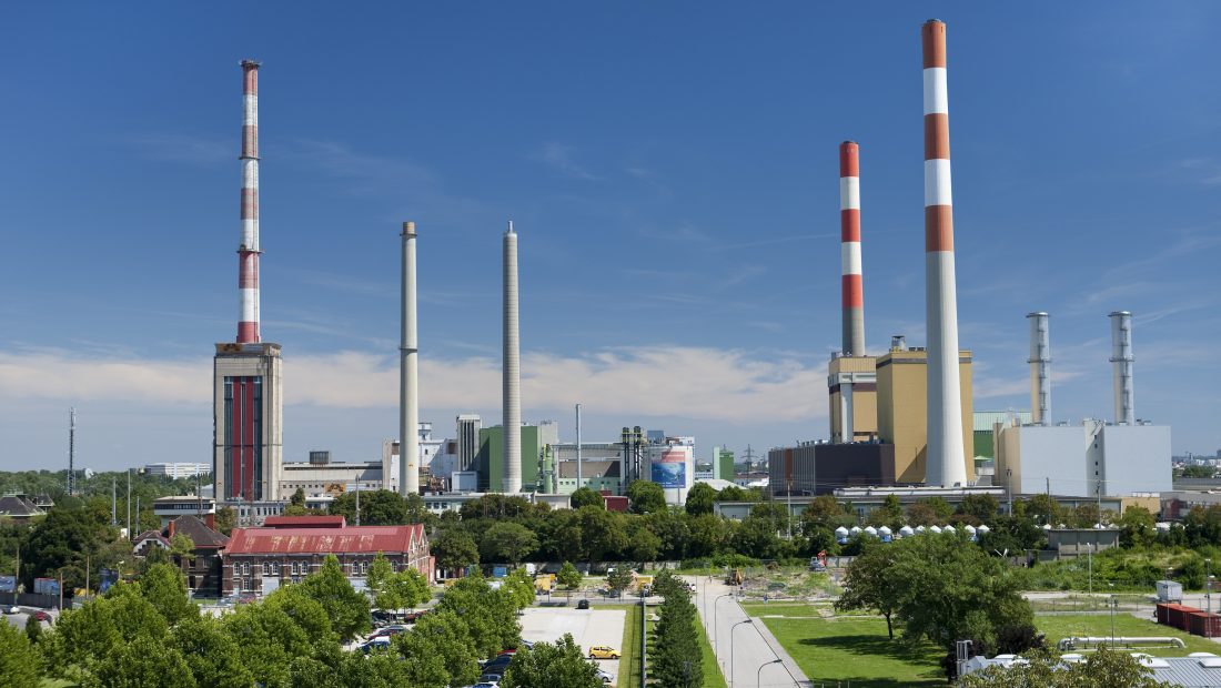 Kraftwerk Simmering mit mehreren Schornsteinen und Gebäuden, umgeben von Grün unter einem blauen Himmel.
