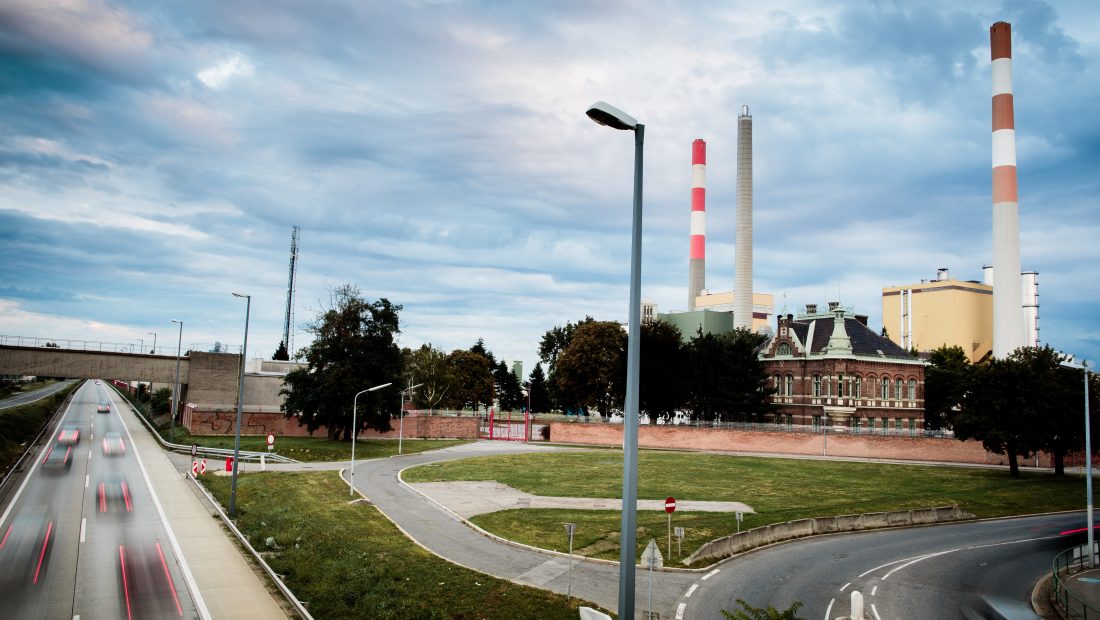 Kraftwerk Simmering mit Schornsteinen in der Nähe einer Autobahn unter bewölktem Himmel. Im Hintergrund Bäume und ein Backsteingebäude.