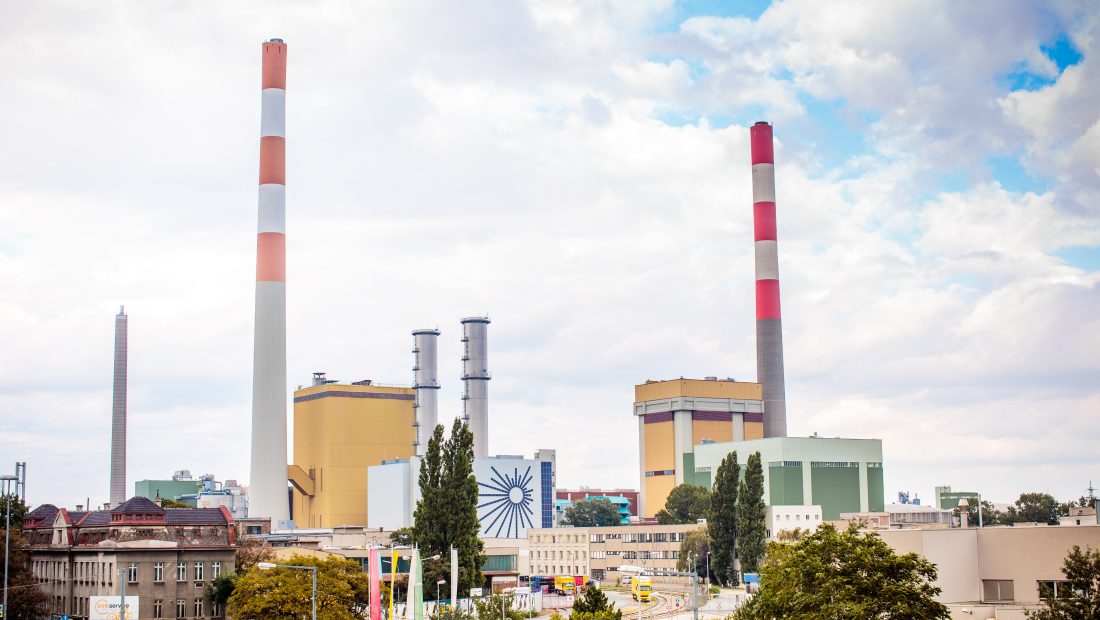Kraftwerk Simmering mit mehreren Schornsteinen unter einem bewölkten Himmel, umgeben von Bäumen und Gebäuden.