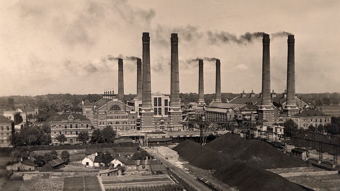 Historisches Foto vom Kraftwerk Simmering mit sechs hohen, Rauch ausstoßenden Schornsteinen, umgeben von Gebäuden und Grünflächen.