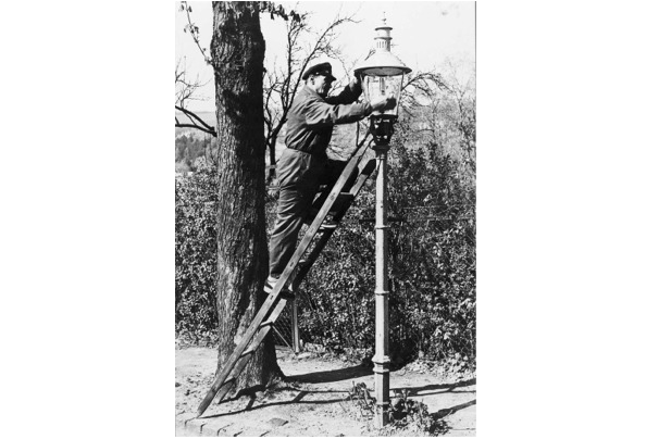 Auf einem Schwarzweißfoto repariert eine Person auf einer Leiter eine Straßenlaterne neben einem Baum.