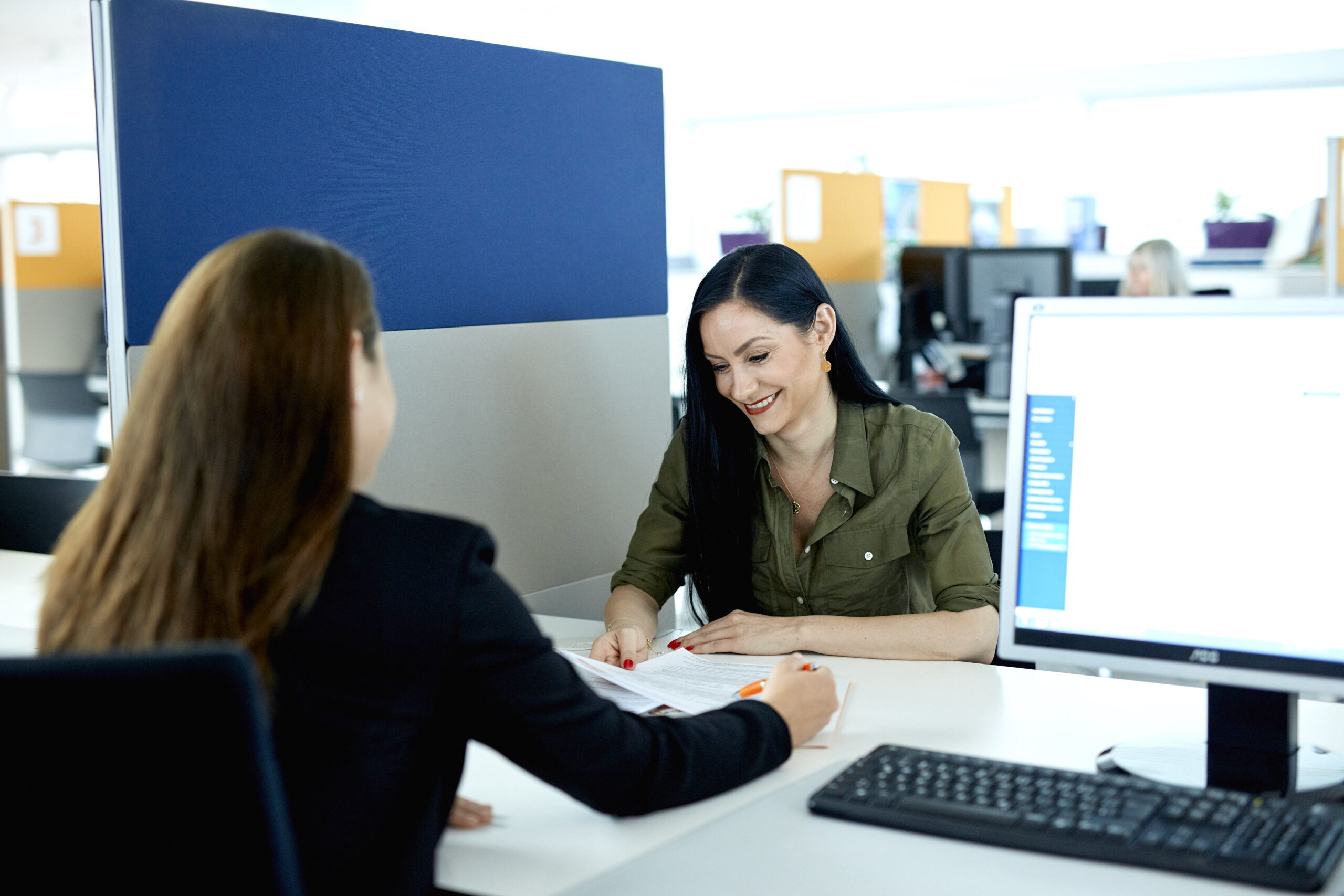 Zwei Frauen sitzen in einem Büro an einem Schreibtisch, lächeln und überprüfen Dokumente auf Papier.