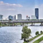 Ein Zug überquert eine Donaubrücke, im Hintergrund ist die Skyline von Wien zu sehen.