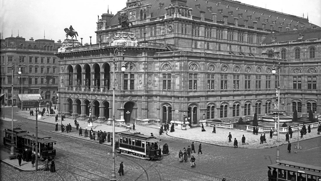 Historische Aufnahme der Wiener Oper mit Straßenbahnen und Menschen im Vordergrund, die auf Kopfsteinpflasterstraßen spazieren.