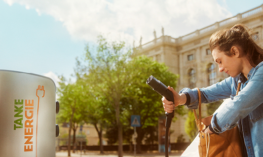 Person, die ein Elektrofahrzeug an einer Ladestation auflädt, im Hintergrund ein Gebäude und Bäume.