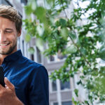 Ein lächelnder Mann im blauen Hemd schaut auf sein Telefon und steht im Freien in der Nähe von Gebäuden und im Grünen.