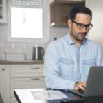 Mann mit Brille arbeitet an einem Laptop an einem Küchentisch, daneben liegen ein Dokument und ein Stift.