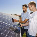 Zwei Personen untersuchen Solarmodule mit Handgeräten unter einem klaren blauen Himmel.