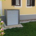 Wärmepumpe außerhalb eines gelben Hauses mit Fensterläden, umgeben von Gras und einem blühenden Busch.