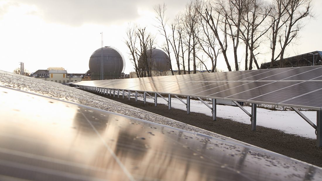 Sonnenkollektoren auf einem schneebedeckten Feld mit Bäumen und zwei großen Industrietanks, dem Kraftwerk Leopoldau, im Hintergrund unter einem bewölkten Himmel.