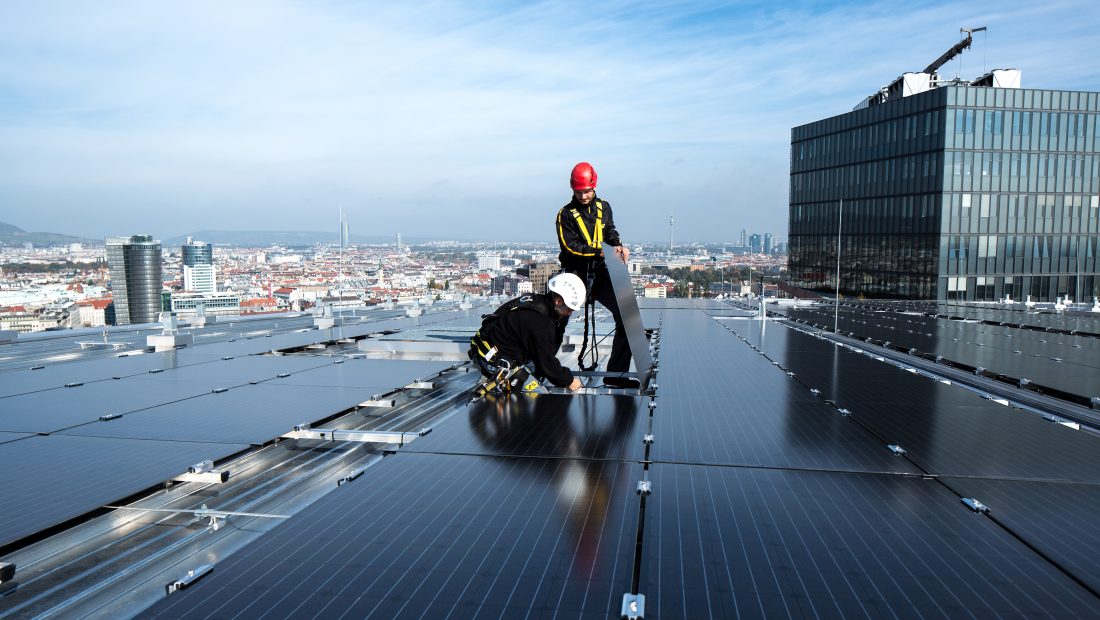 Arbeiter in Sicherheitsausrüstung installieren Solarmodule auf dem Dach The Mall - Wien Mitte, mit der Skyline von Wien im Hintergrund.