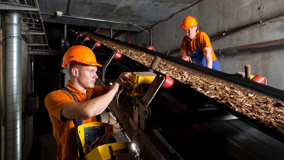 Zwei Arbeiter in orangefarbenen Uniformen und Helmen arbeiten in einer Industrieanlage an einem Förderband, das Holzspäne transportiert.