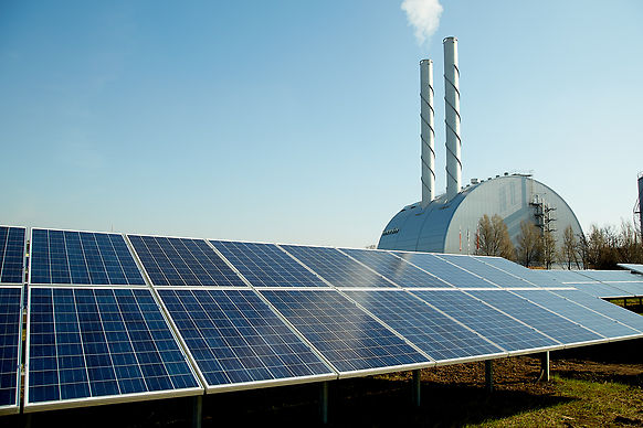 Im Vordergrund ragen markante Solarmodule des BürgerInnen-Solarkraftwerks in den Vordergrund. Dahinter ist das Kraftwerk Inzersdorf bei klarem blauen Himmel zu sehen.