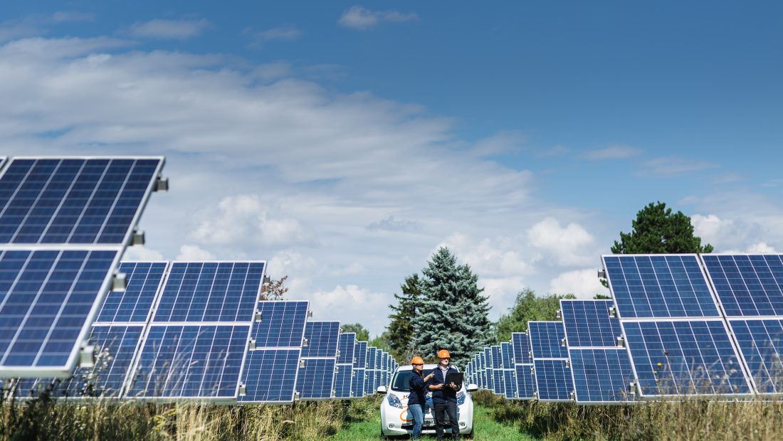 Zwei Techniker mit Helmen inspizieren Solarmodule auf einem Feld unter blauem Himmel.