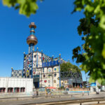 Der Spittelau-Turm und das Spittelau-Gebäude, geschmückt mit einem lebendigen Mosaikmuster, werden von üppigen grünen Blättern unter einem klaren blauen Himmel wunderschön eingerahmt.