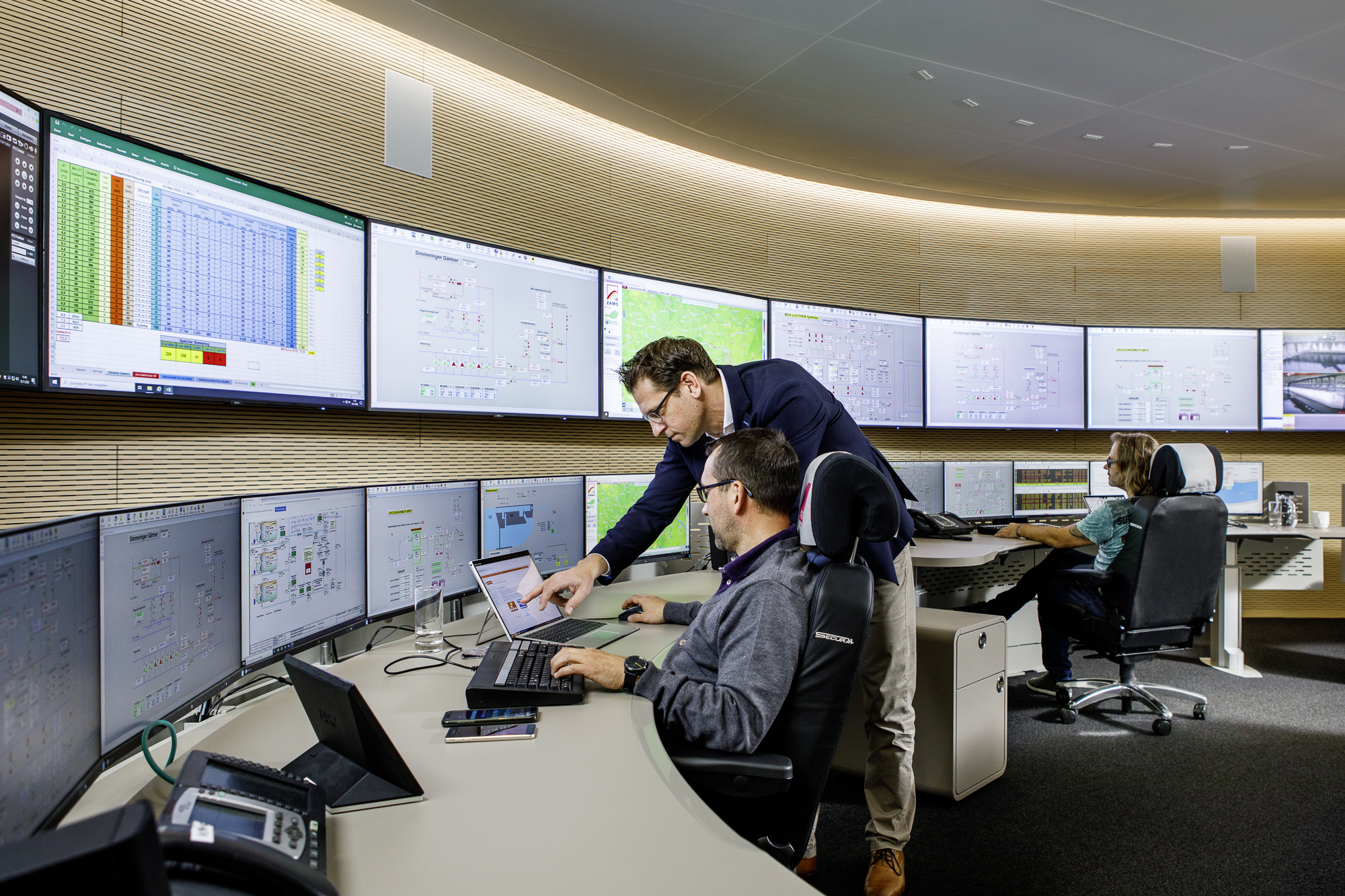 Kontrollzentrum mit vielen Bildschirmen und zwei arbeitetenden Personen