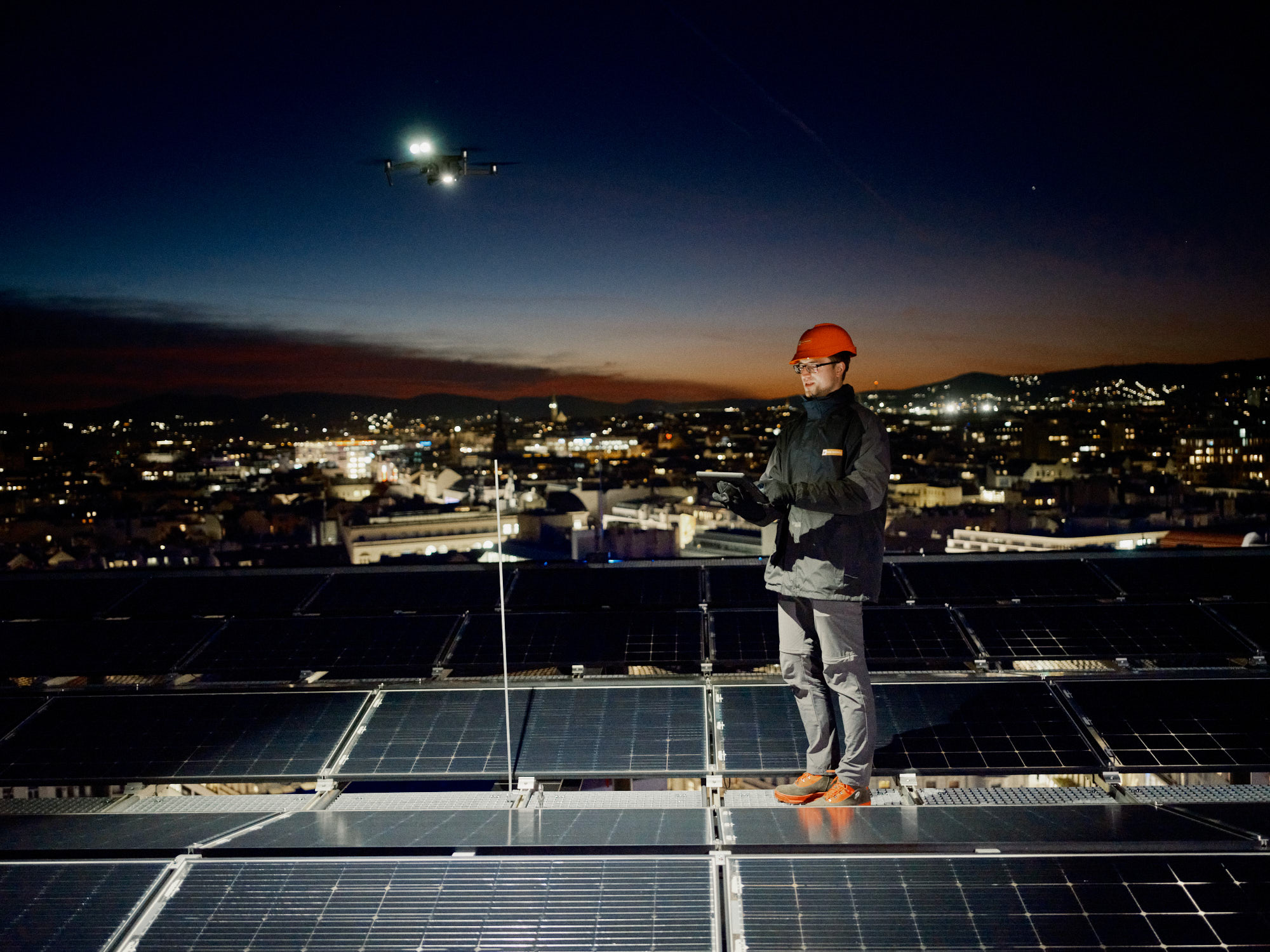 Person mit orangen Wien Energie Helm, die nachts auf Solarmodulen steht und eine Drohne über einer beleuchteten Stadtlandschaft steuert.