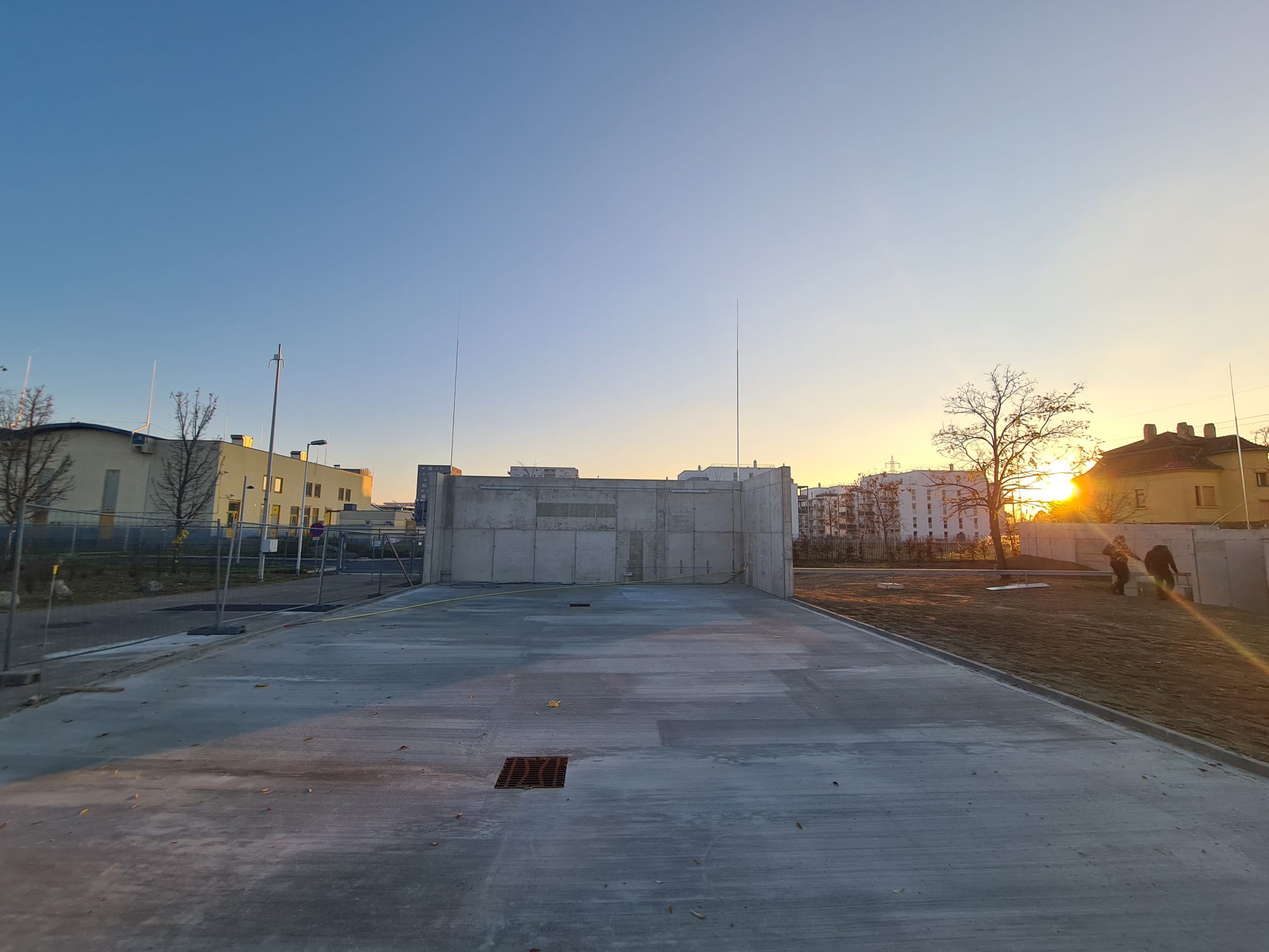 Betonkonstruktion bei Sonnenuntergang, umgeben von Gebäuden und kahlen Bäumen, unter einem klaren Himmel.