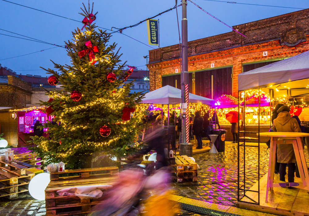Die festlich beleuchtete Adventmarkt-Remise bietet eine bezaubernde Weihnachtsmarktszene mit funkelnden Dekorationen, einem riesigen Weihnachtsbaum, lebhaften Menschen und hellen Ständen.