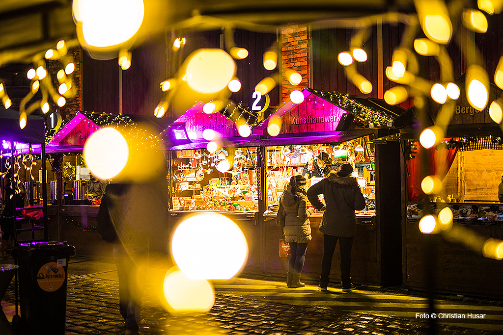 Menschen kaufen auf einem festlichen Adventmarkt im Freien ein, im Vordergrund leuchtende Lichterketten.
