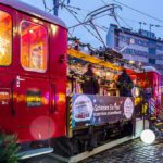 Eine festlich mit Lichtern geschmückte Straßenbahn dient unter dem Abendhimmel als Weihnachtsmarktstand auf einer Stadtstraße.