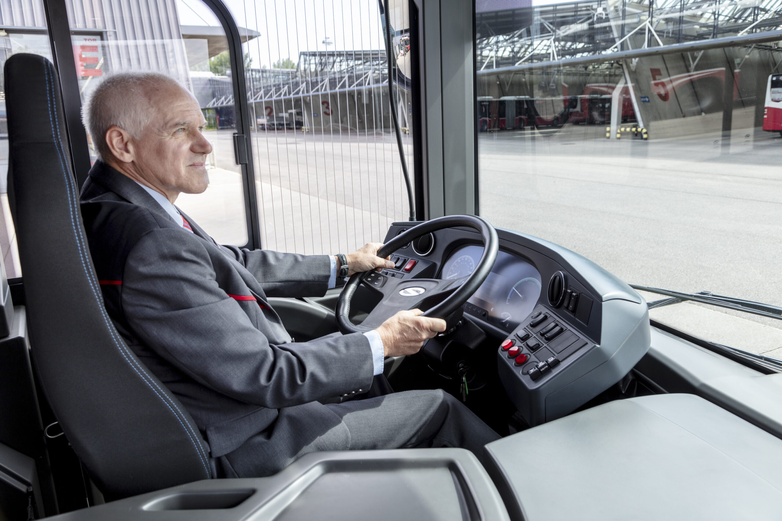 Ein älterer Mann im Anzug fährt einen Bus durch ein Verkehrsgelände.
