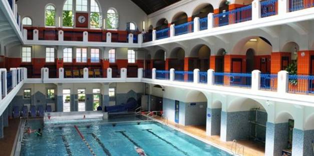 Das Jörgerbad ist ein Hallenbad mit Bahnen, umgeben von Balkonebenen, die mit Bögen und leuchtenden rot-orangen und blauen Akzenten geschmückt sind.