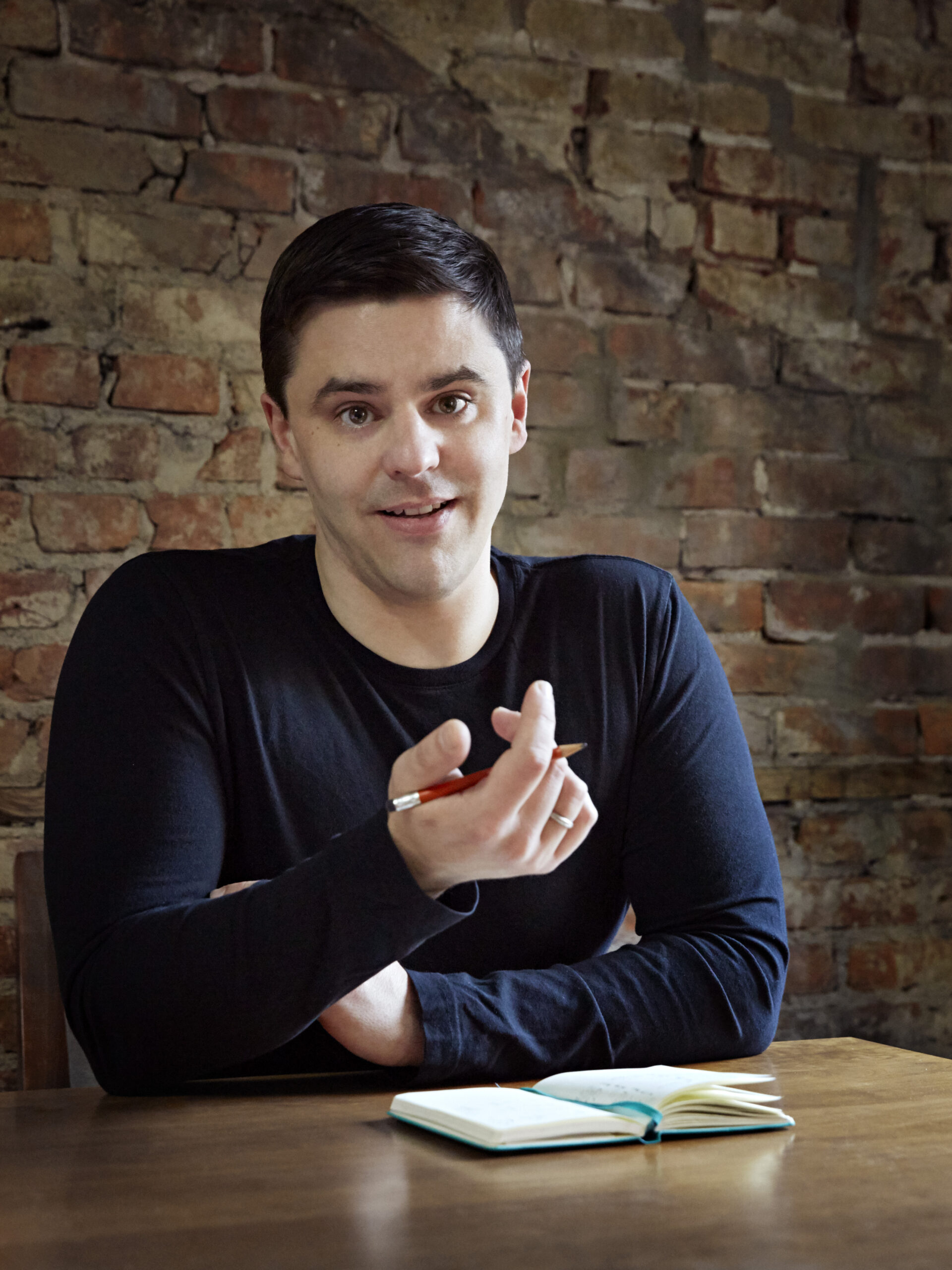 Klimaökonom Gernot in einem dunklen Hemd hält einen Stift und sitzt mit einem Notizbuch an einem Tisch vor der Kulisse hoch aufragender Backsteingebäude.