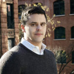 Klimaökonom Gernot Wagner steht im Pullover im Freien, hinter ihm ragen Backsteingebäude auf.