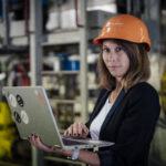 Michaela Kilian trägt einen Bauhelm und konzentriert sich inmitten der geschäftigen Industrieumgebung auf ihren Laptop.