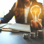 Person, die eine brennende Glühbirne hält und in ein Notizbuch schreibt, auf dem Schreibtisch liegen ein Stapel Münzen und ein Taschenrechner.