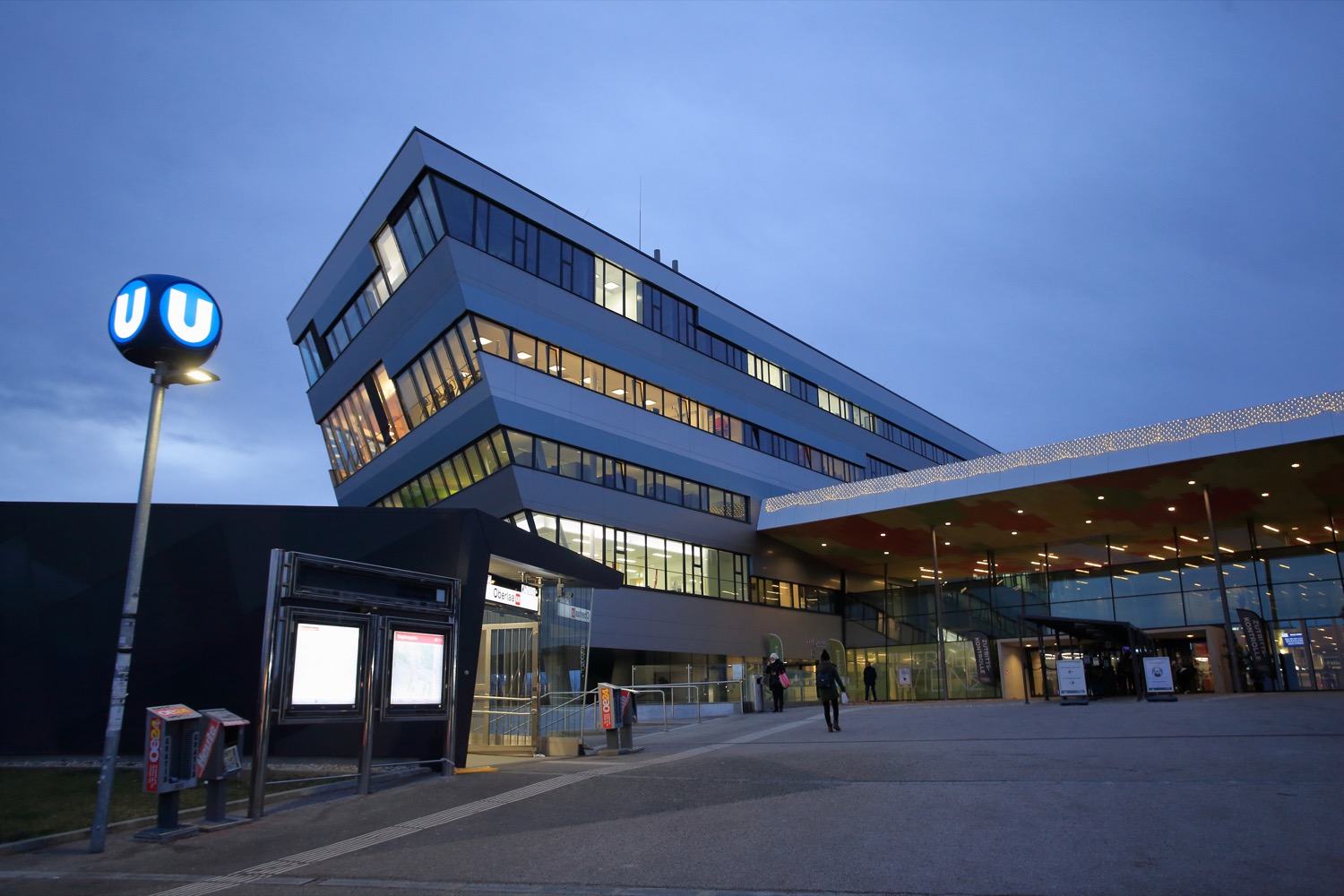Das moderne Gebäude mit seinem kantigen Design und den in der Abenddämmerung beleuchteten Fenstern strahlt eine raffinierte Eleganz aus. Vor der Therme Wien befindet sich die U-Bahn Station Oberlaa.