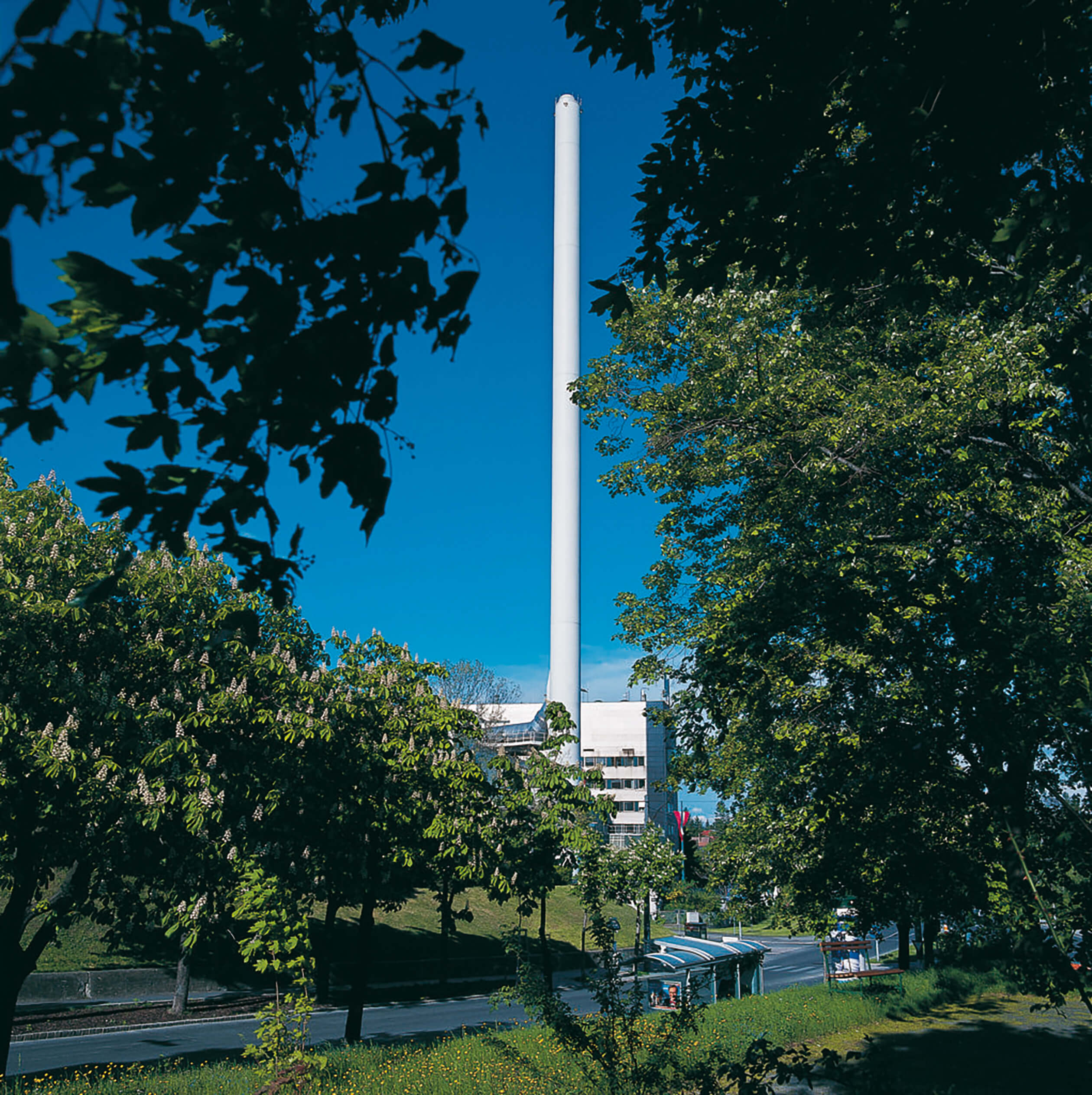 Der hoher, schlanke Schornstein der Abfallverwertungsanlage Flötzersteig erhebt sich über den Gebäuden, umgeben von Bäumen unter einem klaren blauen Himmel.