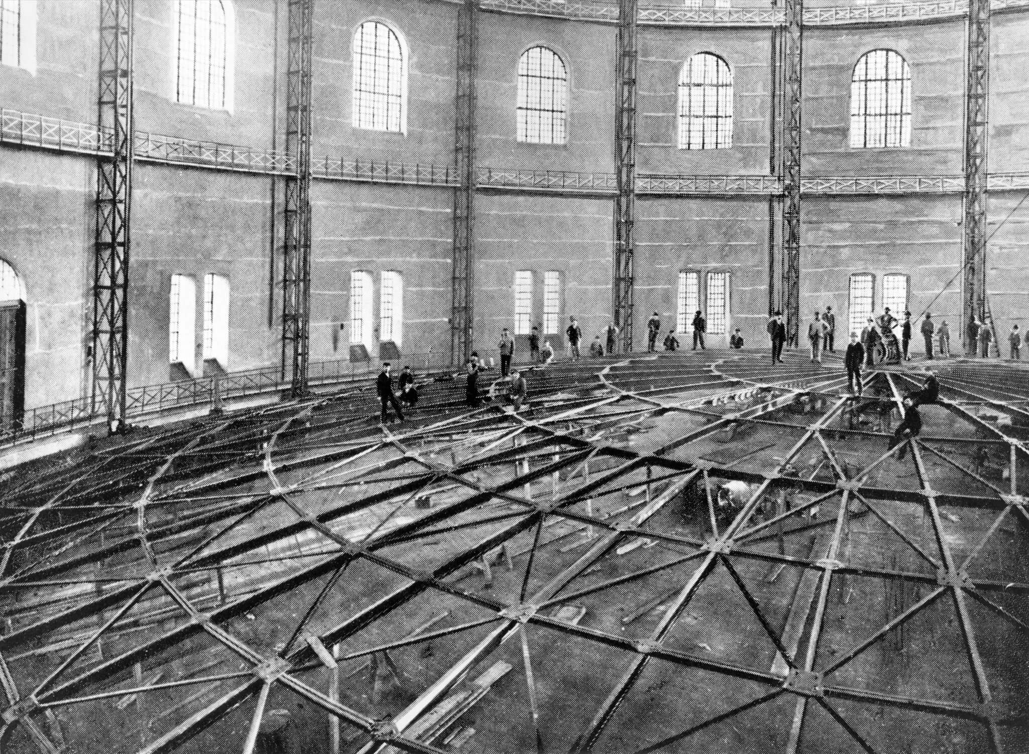 Im großen, runden Gasbehälter mit hohen Fenstern und einem Gerüst montieren Arbeiter ein Metallgerüst. Aufnahme aus dem Jahr 1900.