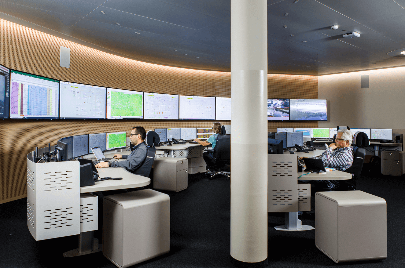 Menschen arbeiten an Computerstationen in einem Kontrollraum mit mehreren großen Bildschirmen an der Wand. Wien Energie Mitarbeiter*innen in der neuen Warte in Erdberg.
