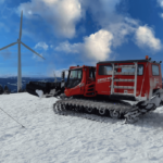 Rote Pistenraupe auf einem schneebedeckten Berg mit einer Windkraftanlage im Hintergrund unter einem blauen Himmel mit Wolken.