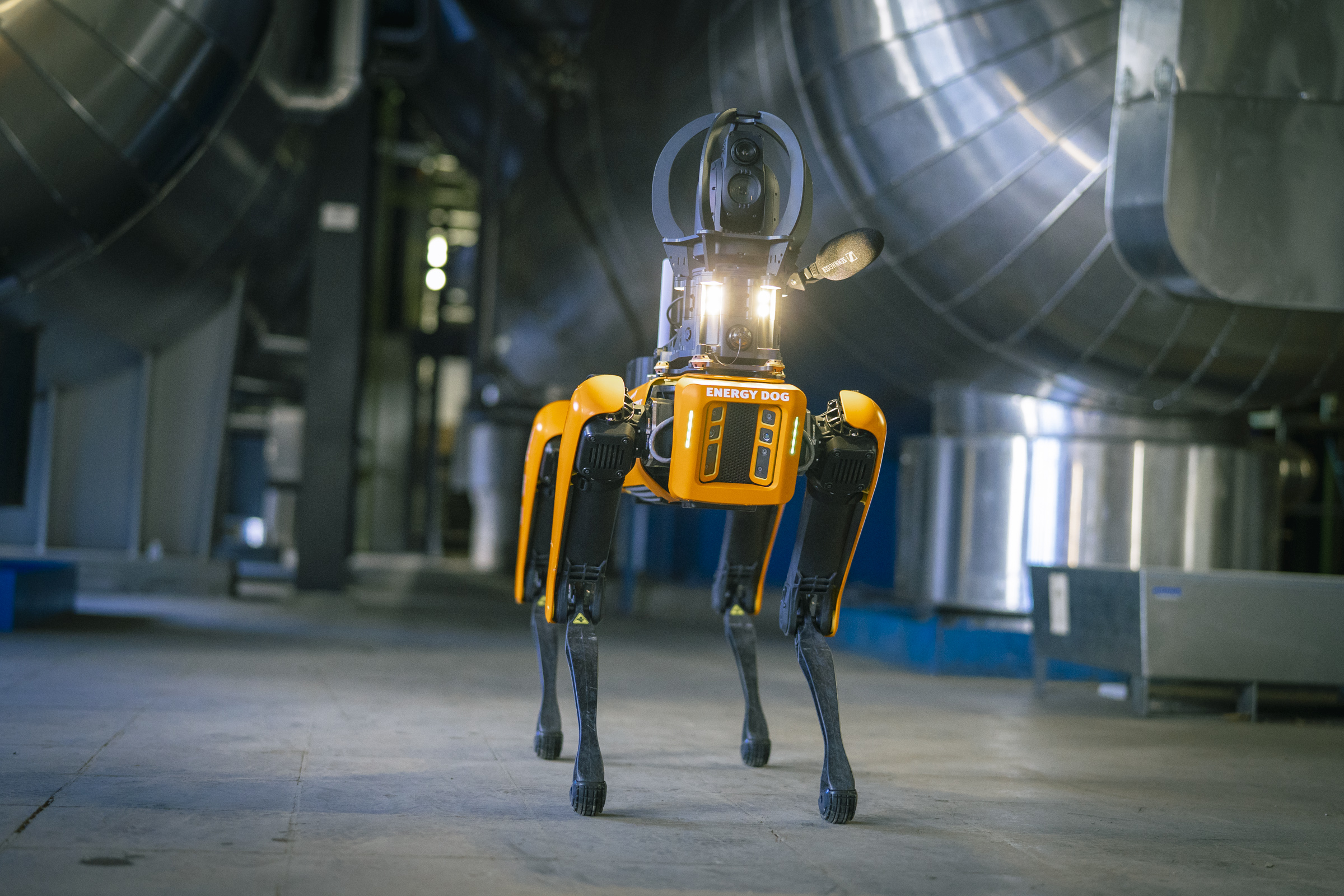 Ein oranger Roboterhund, der "Energy Dog" steht in einer Industrieumgebung mit großen Metallrohren auf dem Boden.