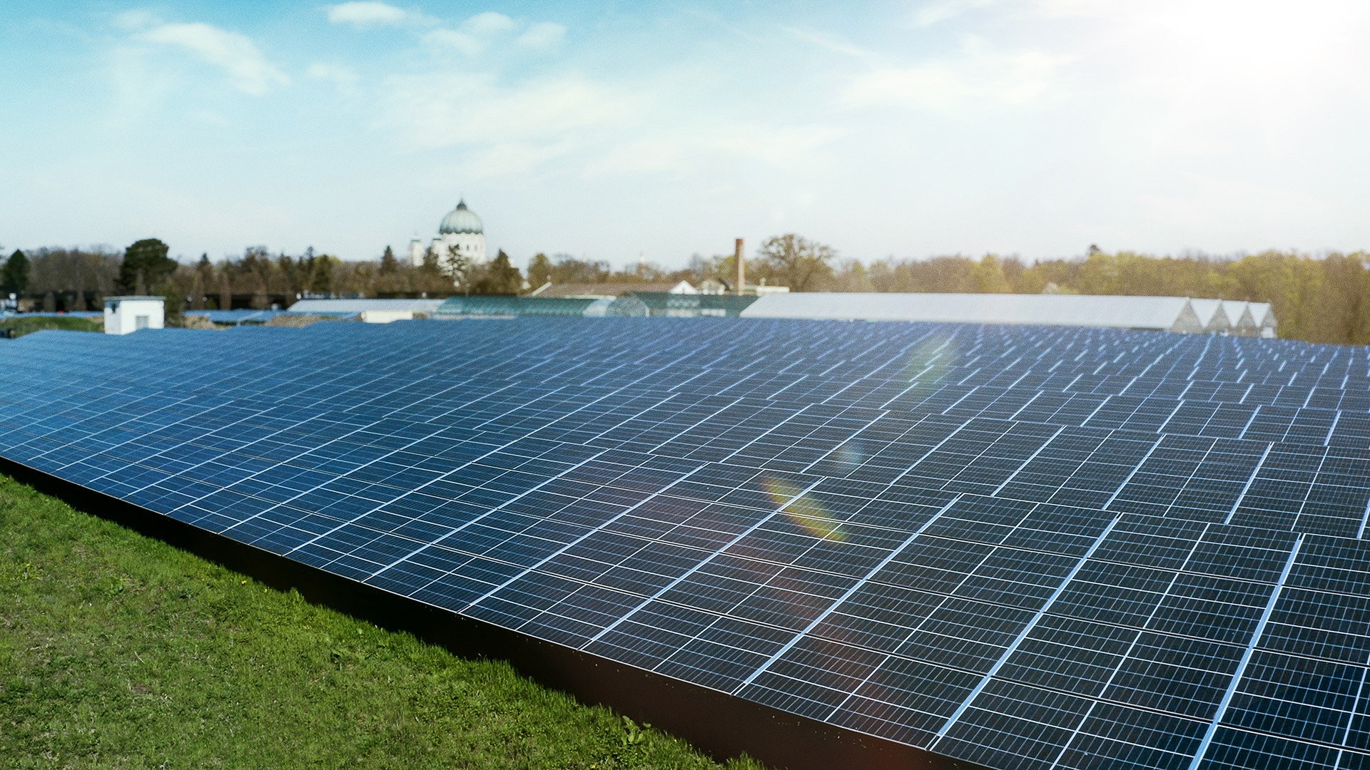 Solarmodule auf dem Gelände des Zentralfriedhofs mit der Friedhofskirche und Bäumen im Hintergrund, alles unter einem klaren blauen Himmel, demonstrieren den innovativen Einsatz einer Photovoltaikanlage.