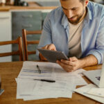 Mann benutzt ein Tablet an einem Tisch mit Papieren, einem Laptop und einer Kanne Kaffee.