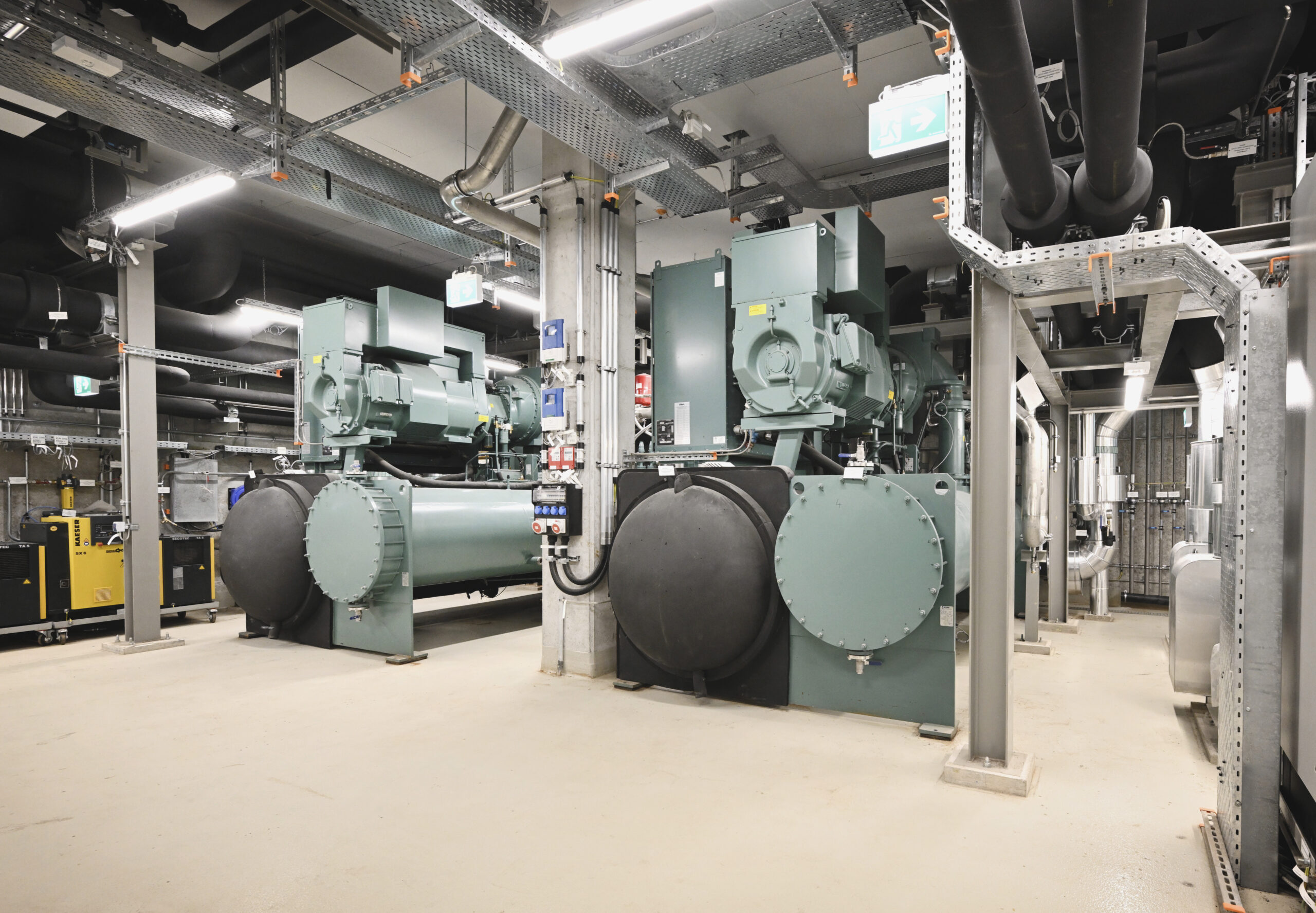Ansicht der angeschlossenen Fernkältezentrale am Stubenring. Industrieraum mit großen grünen Maschinen, Rohren und Kabeln an der Decke in einem gut beleuchteten Raum.