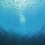 Unterwasseransicht mit durchströmendem Sonnenlicht und Blasen, die in einem tiefblauen Wasser zur Oberfläche aufsteigen.